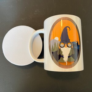 May include: White ceramic mug with a brown oval design featuring a cartoon gnome wearing a blue hat and binoculars. The gnome is in front of a forest scene with a sunset in the background.