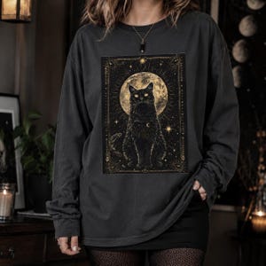 Op de afbeelding: Een antracietgrijs shirt met lange mouwen en een rechthoekige grafische print van een zwarte kat die voor een grote, gedetailleerde volle maan zit. Het ontwerp bevat gouden hemelse elementen, sterren en een decoratieve rand.