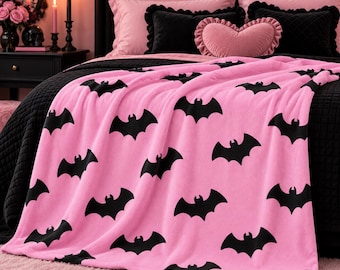 Può includere: Una morbida coperta in pile rosa chiaro con un motivo ripetuto di sagome di pipistrelli neri. La coperta è drappeggiata su un copriletto nero, mettendo in risalto il contrasto tra il tessuto rosa brillante e le forme scure dei pipistrelli.