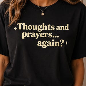 以下が含まれることがあります： 「Thoughts and prayers... again?」の文字がヴィンテージ風のフォントでプリントされた黒いTシャツ。 文字は薄いベージュ色で、小さな星のアクセントが付いています。 シャツはクルーネックです。