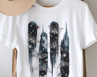 Puede incluir: Camiseta blanca con un diseño de acuarela en blanco y negro de polillas volando frente a un cielo estrellado.