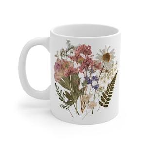 May include: White ceramic mug with a floral design featuring pressed pink, white, and blue flowers. The flowers are arranged in a bouquet shape and are surrounded by green leaves.