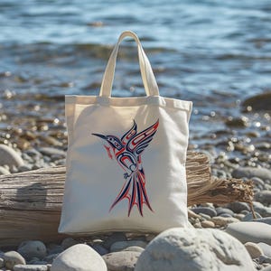 May include: A white canvas tote bag with a red, blue, and black stylized hummingbird design. The bag is sitting on a driftwood log on a beach.