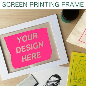May include: A white screen printing frame with a pink insert that reads "YOUR DESIGN HERE." The image also shows printed artwork, ink, and a canvas bag with a printed design. The text "SCREEN PRINTING FRAME" is at the top.