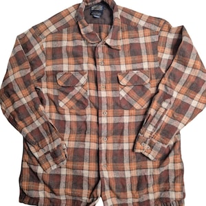 May include: A long-sleeved button-up shirt featuring a brown, tan, and cream plaid pattern. The shirt includes a pointed collar, two chest pockets with buttoned flaps, and a Pendleton brand label visible on the inside of the collar neckline.