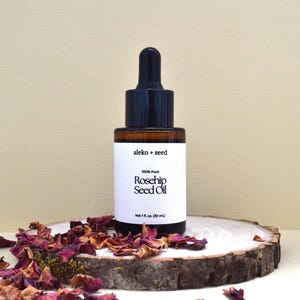 May include: A glass bottle of 100% pure rosehip seed oil with a black dropper top and white label. The label reads "aleko + seed" and "100% Pure Rosehip Seed Oil" with a net volume of 1 fl. oz. (30 mL). The bottle is sitting on a wooden slice with dried rose petals.