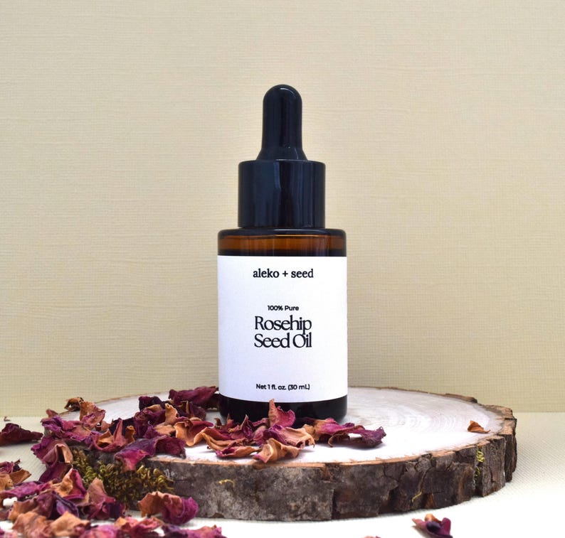 May include: A glass bottle of 100% pure rosehip seed oil with a black dropper top and white label. The label reads "aleko + seed" and "100% Pure Rosehip Seed Oil" with a net volume of 1 fl. oz. (30 mL). The bottle is sitting on a wooden slice with dried rose petals.