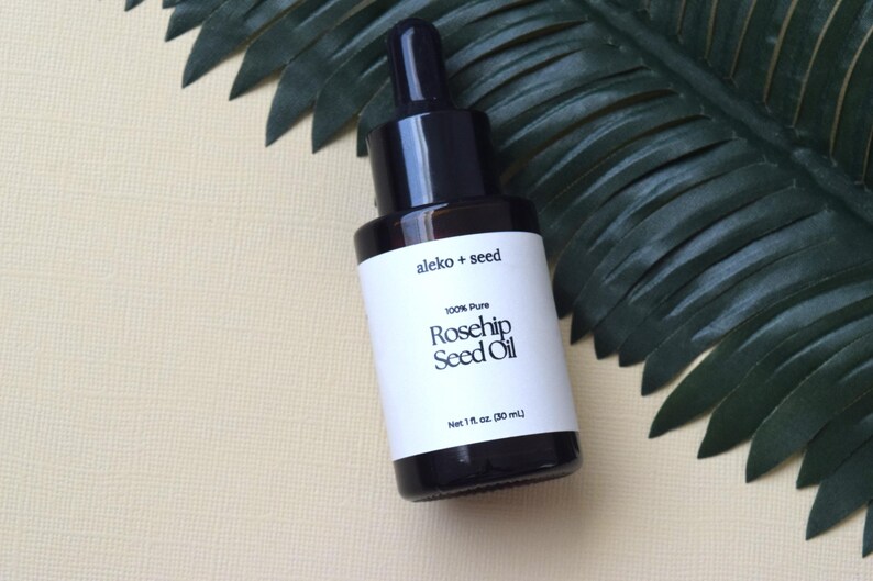 May include: A glass bottle with a black dropper top and a white label. The label says "aleko + seed 100% Pure Rosehip Seed Oil Net 1 fl. oz. (30 ml)"