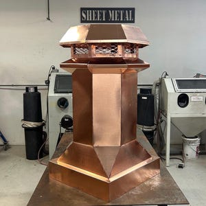 May include: A copper chimney cap with a square base and a hexagonal top. The cap has a wire mesh screen on the top and the words "SHEET METAL" are visible on a sign in the background.