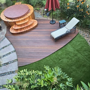 May include: An outdoor living space featuring a wooden hot tub with steps, a lounge chair, and a red umbrella. The deck is made of dark wood, and there is a patch of green artificial turf. Lush greenery surrounds the area.