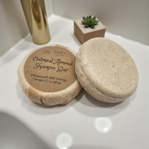 May include: Two round oatmeal almond shampoo bars. One is wrapped in brown paper with the text "Oatmeal Almond Shampoo Bar" and "Infused with Reiki Energy, Weight 2.2 oz/65 gr". The other bar is unwrapped and light brown.