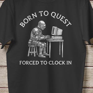 May include: Black t-shirt with a graphic of a knight in armor sitting at a computer. The text reads "BORN TO QUEST" above the knight and "FORCED TO CLOCK IN" below. The shirt is hanging on a wooden background.