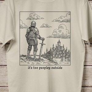 May include: Beige t-shirt with a black and white illustration of a knight standing on a hill, holding a sword, with a castle in the background. The text "it's too peopley outside" is printed below the illustration.