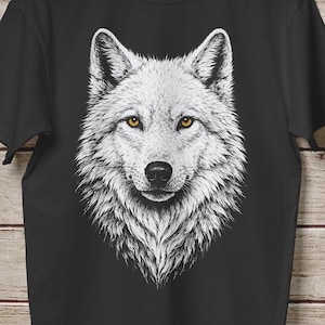 May include: Black t-shirt featuring a detailed white wolf illustration. The wolf's face is centered, with yellow eyes and a detailed fur texture. The shirt is hanging on a wooden hanger against a wood-paneled background.