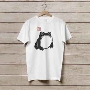 K&ouml;nnte beinhalten: Wei&szlig;es T-Shirt mit einer schwarzen Frosch-Grafik, die einen gro&szlig;en offenen Mund und zwei Augen zeigt. &Uuml;ber dem Frosch befindet sich ein rotes stempelartiges Design. Das T-Shirt h&auml;ngt an einem Holzb&uuml;gel vor einem Holzpaneel-Hintergrund.