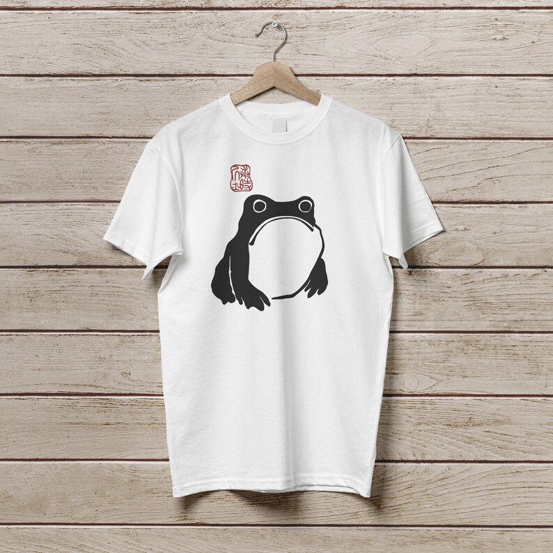 K&ouml;nnte beinhalten: Wei&szlig;es T-Shirt mit einer schwarzen Frosch-Grafik, die einen gro&szlig;en offenen Mund und zwei Augen zeigt. &Uuml;ber dem Frosch befindet sich ein rotes stempelartiges Design. Das T-Shirt h&auml;ngt an einem Holzb&uuml;gel vor einem Holzpaneel-Hintergrund.