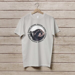 K&ouml;nnte beinhalten: Ein wei&szlig;es T-Shirt mit einer kreisf&ouml;rmigen Grafik eines Waschb&auml;ren mit dem Wort "PEDRO" wiederholt um den Kreis herum.