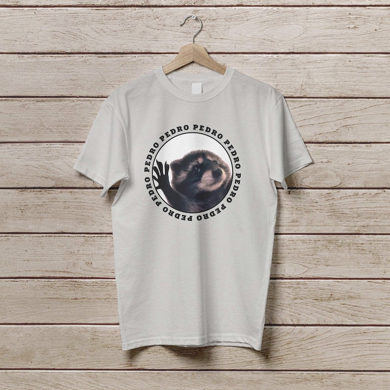 K&ouml;nnte beinhalten: Ein wei&szlig;es T-Shirt mit einer kreisf&ouml;rmigen Grafik eines Waschb&auml;ren mit dem Wort "PEDRO" wiederholt um den Kreis herum.