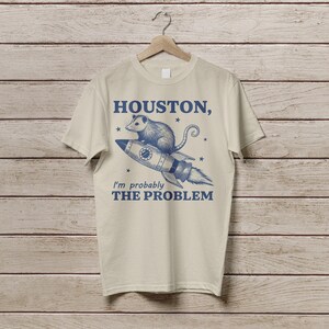 Może przedstawiać: Beżowa koszulka z niebieskim nadrukiem przedstawiającym oposa na rakiecie z tekstem "HOUSTON, I'm probably THE PROBLEM".