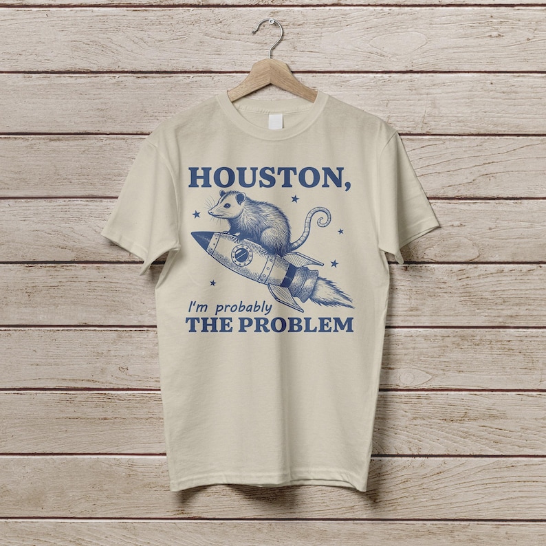 Może przedstawiać: Beżowa koszulka z niebieskim nadrukiem przedstawiającym oposa na rakiecie z tekstem "HOUSTON, I'm probably THE PROBLEM".