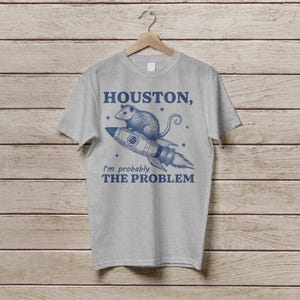 Może przedstawiać: Szary T-shirt z niebieskim nadrukiem przedstawiającym oposa na rakiecie z tekstem "Houston, prawdopodobnie jestem problemem".