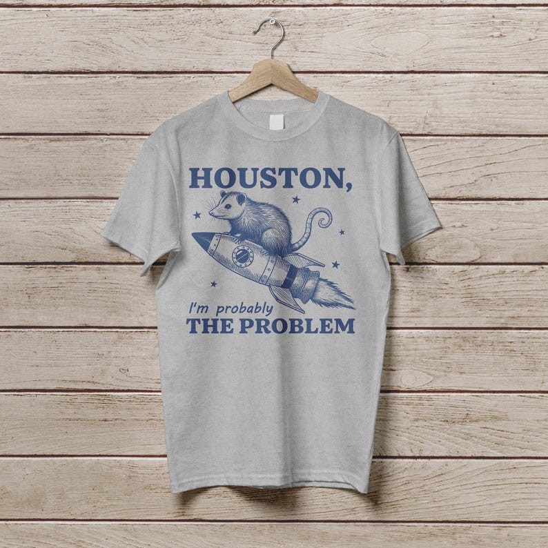 Może przedstawiać: Szary T-shirt z niebieskim nadrukiem przedstawiającym oposa na rakiecie z tekstem "Houston, prawdopodobnie jestem problemem".