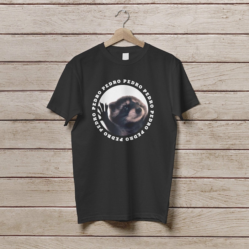 K&ouml;nnte beinhalten: Schwarzes T-Shirt mit einem wei&szlig;en Kreis-Motiv, das einen Waschb&auml;ren und den Text "PEDRO" zeigt, der sich um den Kreis wiederholt.