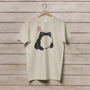 K&ouml;nnte beinhalten: Beiges T-Shirt mit einem schwarzen Frosch-Motiv. Der Frosch hat einen gro&szlig;en, offenen Mund und zwei runde Augen. &Uuml;ber dem Frosch befindet sich ein rotes, stempelartiges Design. Das T-Shirt h&auml;ngt an einem Holzb&uuml;gel.