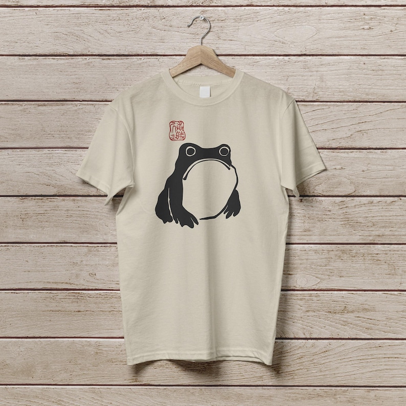 K&ouml;nnte beinhalten: Beiges T-Shirt mit einem schwarzen Frosch-Motiv. Der Frosch hat einen gro&szlig;en, offenen Mund und zwei runde Augen. &Uuml;ber dem Frosch befindet sich ein rotes, stempelartiges Design. Das T-Shirt h&auml;ngt an einem Holzb&uuml;gel.