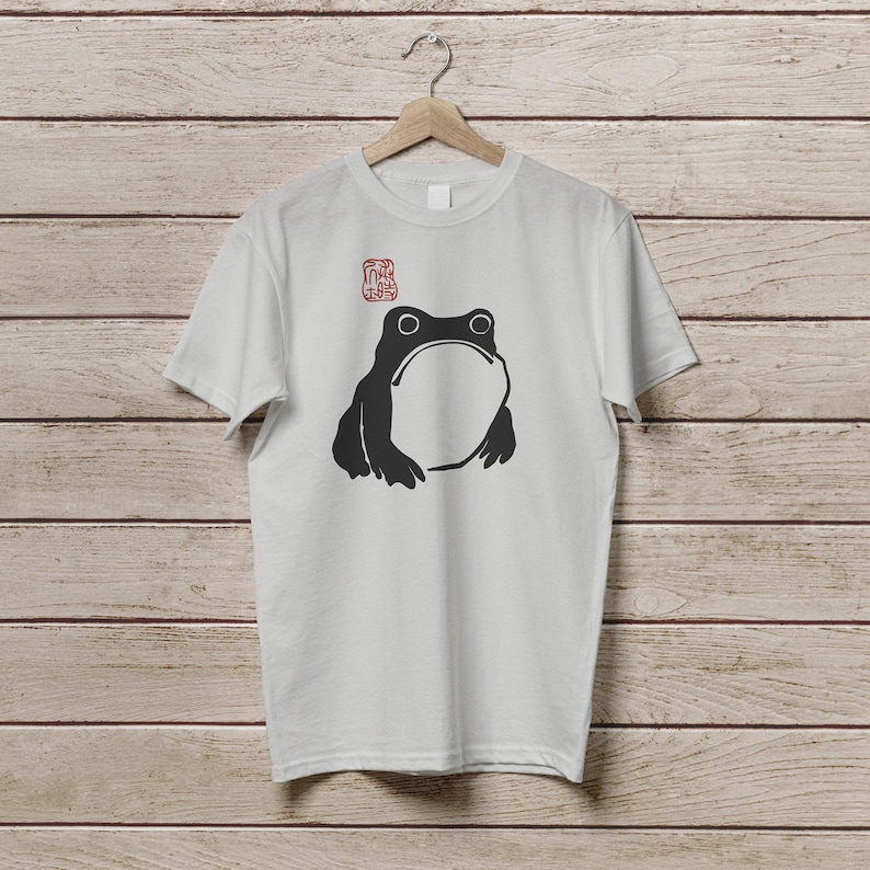 K&ouml;nnte beinhalten: Wei&szlig;es T-Shirt mit einer schwarzen Frosch-Illustration, die einen gro&szlig;en offenen Mund und zwei Augen zeigt. &Uuml;ber dem Frosch befindet sich ein rotes stempelartiges Design. Das Shirt h&auml;ngt an einem Holzb&uuml;gel vor einem Holzpaneel-Hintergrund.