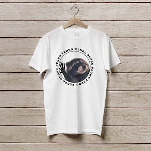 K&ouml;nnte beinhalten: Wei&szlig;es T-Shirt mit einem schwarzen Kreis-Motiv, das einen Waschb&auml;ren mit dem Wort "PEDRO" zeigt, das sich um den Kreis wiederholt.