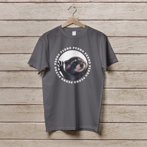 K&ouml;nnte beinhalten: Ein dunkelgraues T-Shirt mit einem kreisf&ouml;rmigen Grafikdruck eines Waschb&auml;ren mit dem Wort "PEDRO" wiederholt um den Kreis herum.