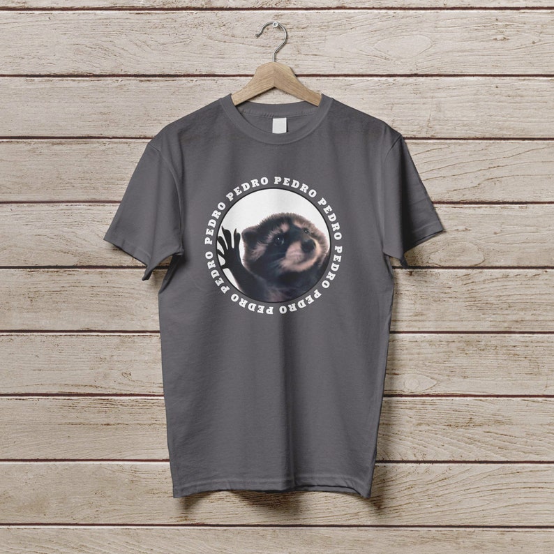 K&ouml;nnte beinhalten: Ein dunkelgraues T-Shirt mit einem kreisf&ouml;rmigen Grafikdruck eines Waschb&auml;ren mit dem Wort "PEDRO" wiederholt um den Kreis herum.