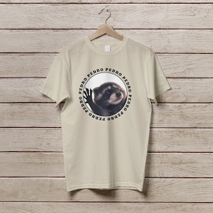 K&ouml;nnte beinhalten: Ein beiges T-Shirt mit einem kreisf&ouml;rmigen Grafikdruck eines Waschb&auml;ren, um den Kreis herum steht der Name "PEDRO" wiederholt.