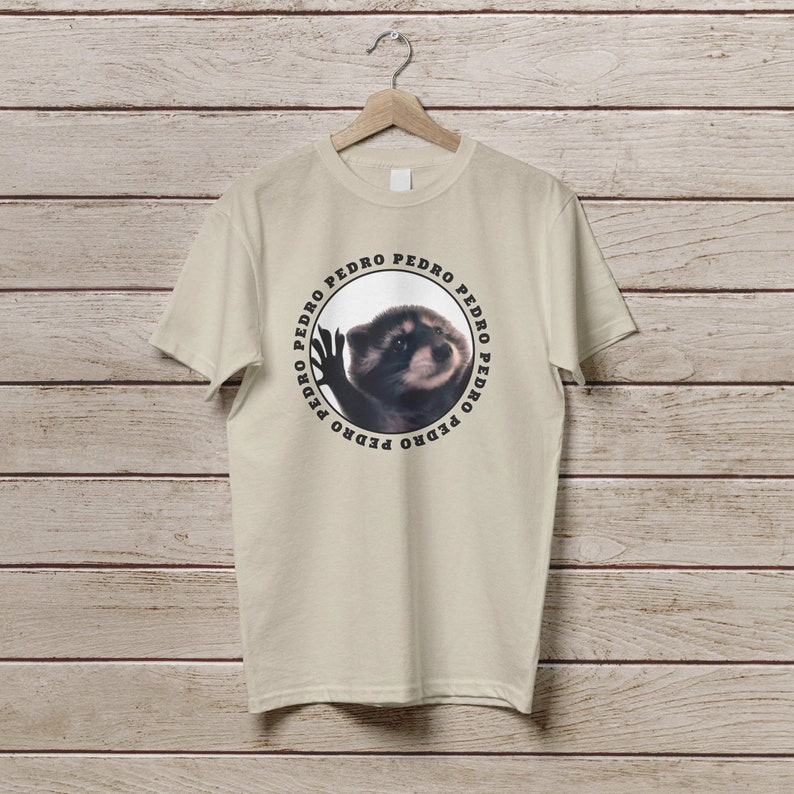 K&ouml;nnte beinhalten: Ein beiges T-Shirt mit einem kreisf&ouml;rmigen Grafikdruck eines Waschb&auml;ren, um den Kreis herum steht der Name "PEDRO" wiederholt.