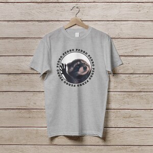 K&ouml;nnte beinhalten: Ein graues T-Shirt mit einem runden Design, das einen Waschb&auml;ren mit dem Wort "PEDRO" zeigt, das um den Kreis herum wiederholt wird.