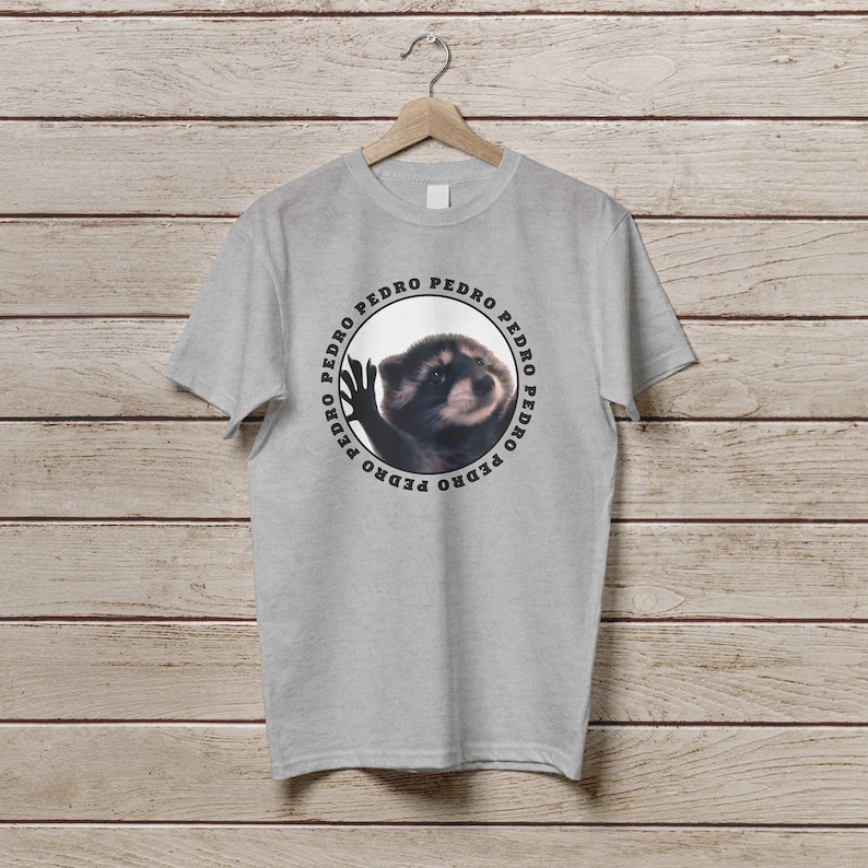 K&ouml;nnte beinhalten: Ein graues T-Shirt mit einem runden Design, das einen Waschb&auml;ren mit dem Wort "PEDRO" zeigt, das um den Kreis herum wiederholt wird.
