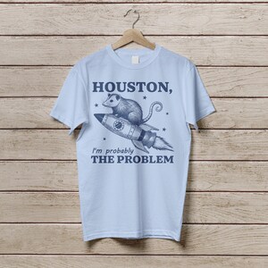Może przedstawiać: Jasnoniebieska koszulka z nadrukiem oposa jadącego rakietą z tekstem "Houston, I'm probably the problem" nadrukowanym z przodu.