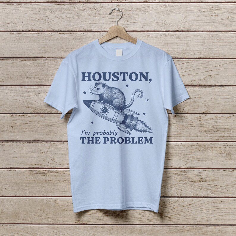 Może przedstawiać: Jasnoniebieska koszulka z nadrukiem oposa jadącego rakietą z tekstem "Houston, I'm probably the problem" nadrukowanym z przodu.