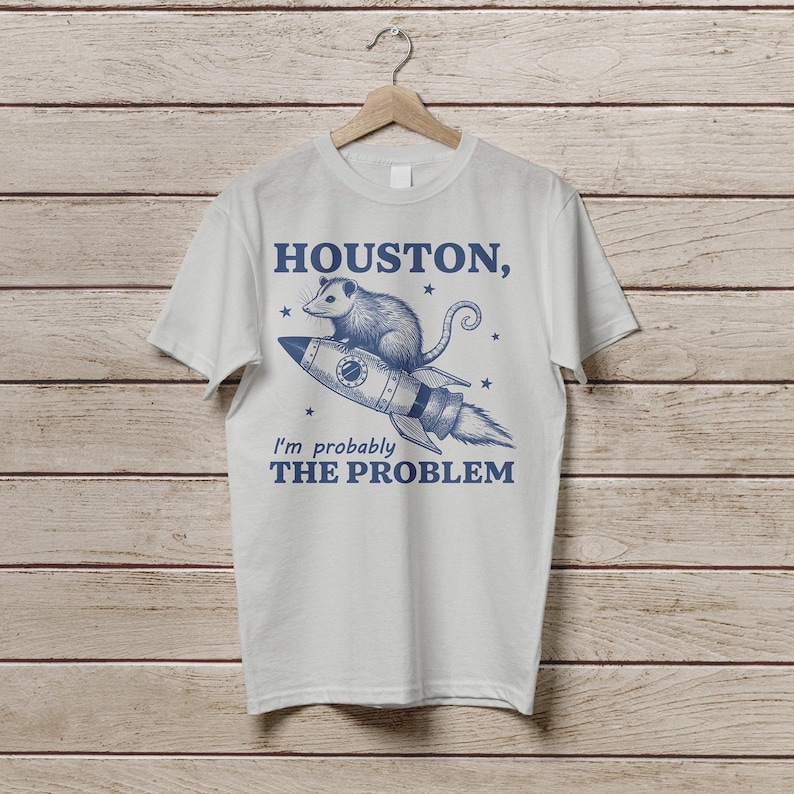 Może przedstawiać: Biały t-shirt z niebieskim nadrukiem oposa jadącego rakietą z tekstem "HOUSTON, I'm probably THE PROBLEM" nadrukowanym na nim.