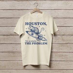 Może przedstawiać: Kremowa koszulka z niebieskim nadrukiem przedstawiającym oposa na rakiecie z tekstem "HOUSTON, I'm probably THE PROBLEM" nadrukowanym z przodu.
