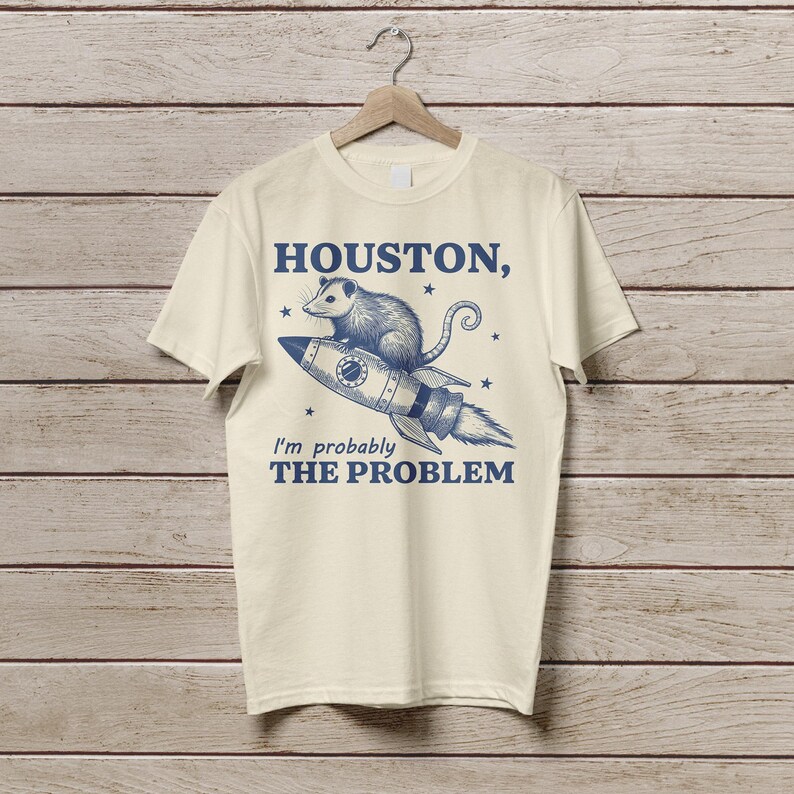 Może przedstawiać: Kremowa koszulka z niebieskim nadrukiem przedstawiającym oposa na rakiecie z tekstem "HOUSTON, I'm probably THE PROBLEM" nadrukowanym z przodu.