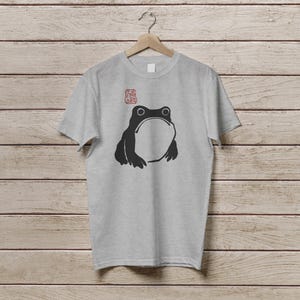 K&ouml;nnte beinhalten: Ein hellgraues T-Shirt mit einer schwarzen Frosch-Illustration, die einen gro&szlig;en offenen Mund und zwei runde Augen zeigt. &Uuml;ber dem Frosch befindet sich ein roter Stempel. Das T-Shirt h&auml;ngt an einem Holzb&uuml;gel.