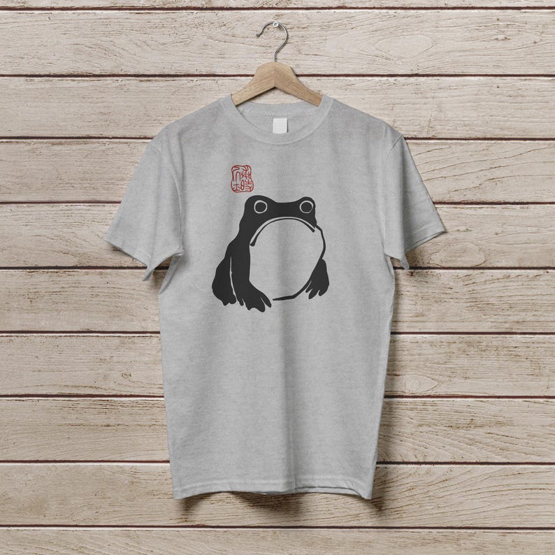 K&ouml;nnte beinhalten: Ein hellgraues T-Shirt mit einer schwarzen Frosch-Illustration, die einen gro&szlig;en offenen Mund und zwei runde Augen zeigt. &Uuml;ber dem Frosch befindet sich ein roter Stempel. Das T-Shirt h&auml;ngt an einem Holzb&uuml;gel.