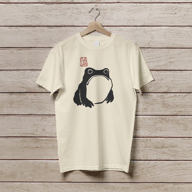 K&ouml;nnte beinhalten: Cremefarbenes T-Shirt mit einer schwarzen Frosch-Illustration. Der Frosch hat einen gro&szlig;en offenen Mund und zwei runde Augen. Oberhalb des Frosches befindet sich ein rotes stempelartiges Design. Das T-Shirt h&auml;ngt an einem Holzkleiderb&uuml;gel.