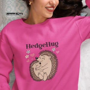 Può includere: Una felpa rosa acceso con la scritta "HedgeHug" sopra un'illustrazione di due ricci che si abbracciano. La felpa è a tinta unita con girocollo e maniche lunghe. L'immagine include anche il testo "Lemonade Pink".