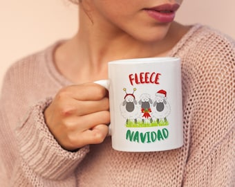 Könnte beinhalten: Weiße Keramiktasse mit drei Cartoon-Schafen mit festlicher Dekoration. Auf der Tasse steht das Wort "FLEECE" in Rot und "NAVIDAD" in Grün. Die Tasse wird von einer Person in einem rosa Pullover gehalten.