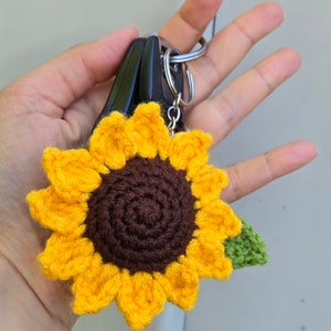 May include: A crocheted sunflower keyring with a brown centre and green leaves. The keyring is attached to a silver key ring.