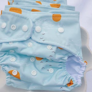 May include: A light blue cloth nappy with a white and yellow sun and bird print. The nappy has white poppers and a white elastic waistband.
