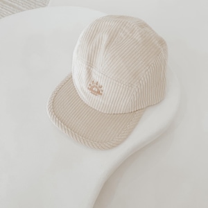 May include: A beige corduroy baseball cap with a small embroidered sun design. The cap has a flat brim and a structured crown.